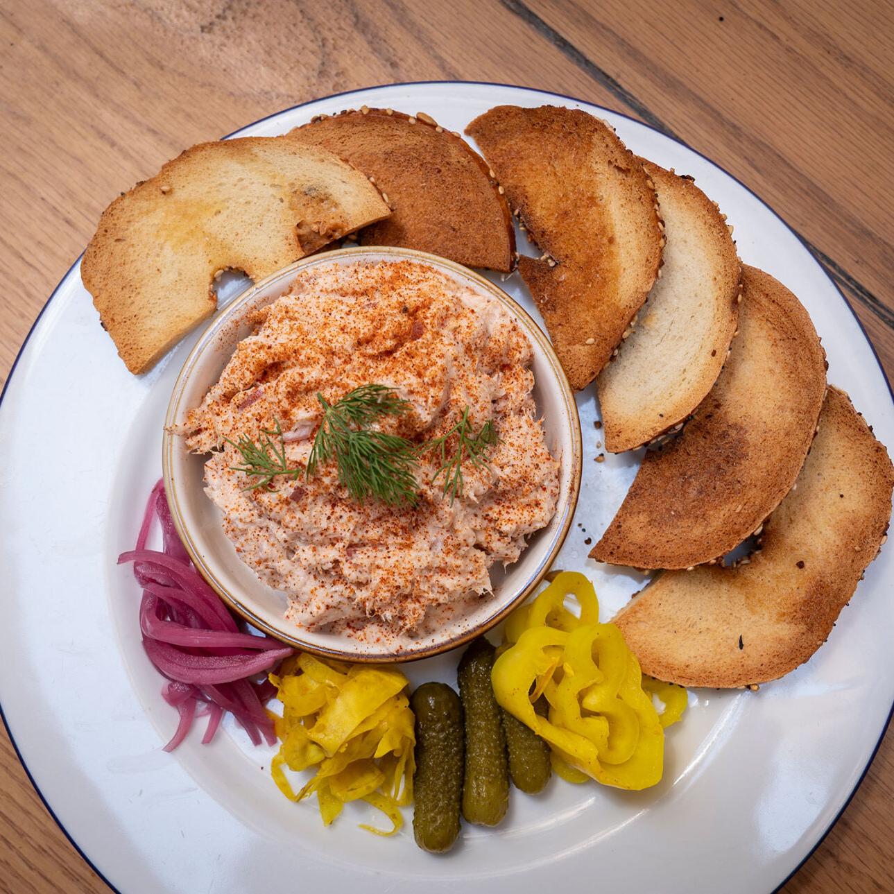 A bowl of fish dip on a plate surrounded by slices of toasted bread and pickled vegetables